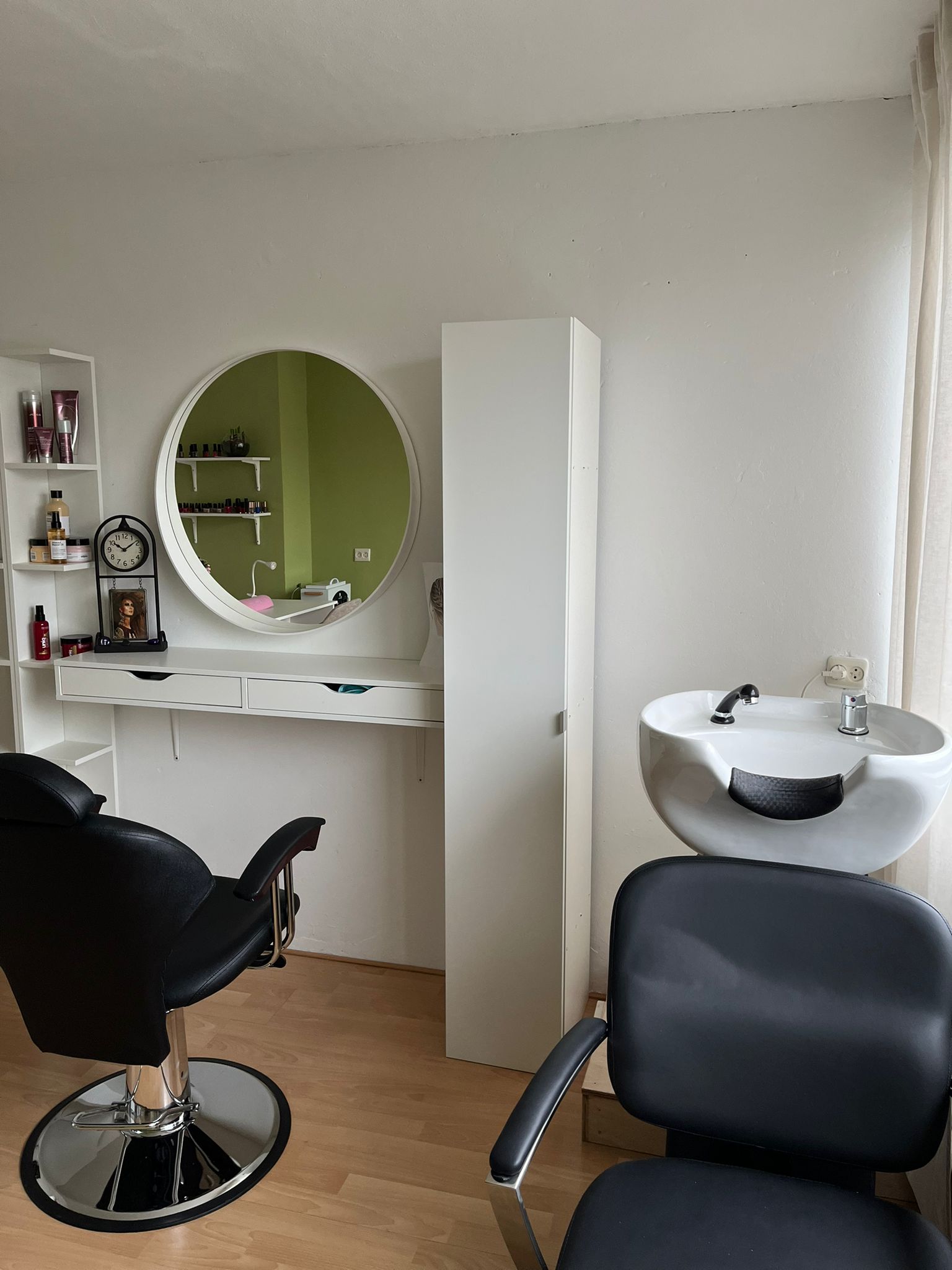 De kappershoek van de salon: ronde spiegel, zwarte kappersstoel en een witte wasbak