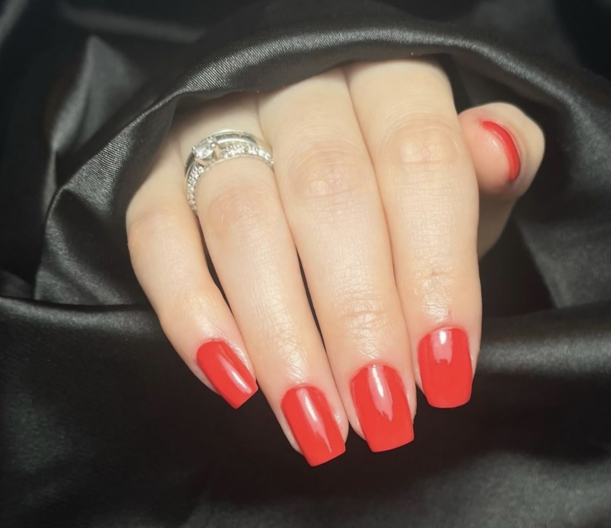 Rode vierkante gelnagels met een ring, op zwart satijn, gedaan bij Manu's Beauty Salon
